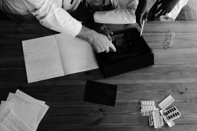 Profissionais da saúde colaboram na análise de dados médicos usando tablets e cadernos em um ambiente monocromático.