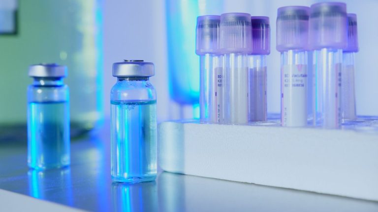 Frascos e tubos de ensaio com líquidos azuis em um laboratório sob luz fluorescente.